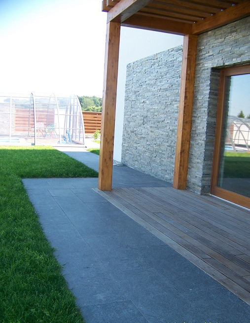 Nowoczesny taras z desek kompozytowych przechodzący w ścieżkę z ciemnej kostki brukowej, z drewnianą pergolą i kamienną ścianą domu w tle, widoczny basen z zadaszeniem.