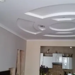 Sufit podwieszany z dekoracyjnymi elementami gipsowymi w kształcie okręgów i kwadratów, oświetlenie punktowe, ściana akcentowa z kamienia dekoracyjnego, widok na kuchnię z białymi szafkami.