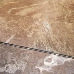 Ułożone brązowe płytki podłogowe imitujące kamień z metalową listwą dylatacyjną wzdłuż krawędzi ściany.