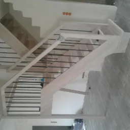 Wnętrze z surową konstrukcją schodów z jasnego drewna i czarnymi, poziomymi balustradami, widoczne elementy instalacji elektrycznej na ścianie, w tle fragment schodów z drewnianymi stopniami.