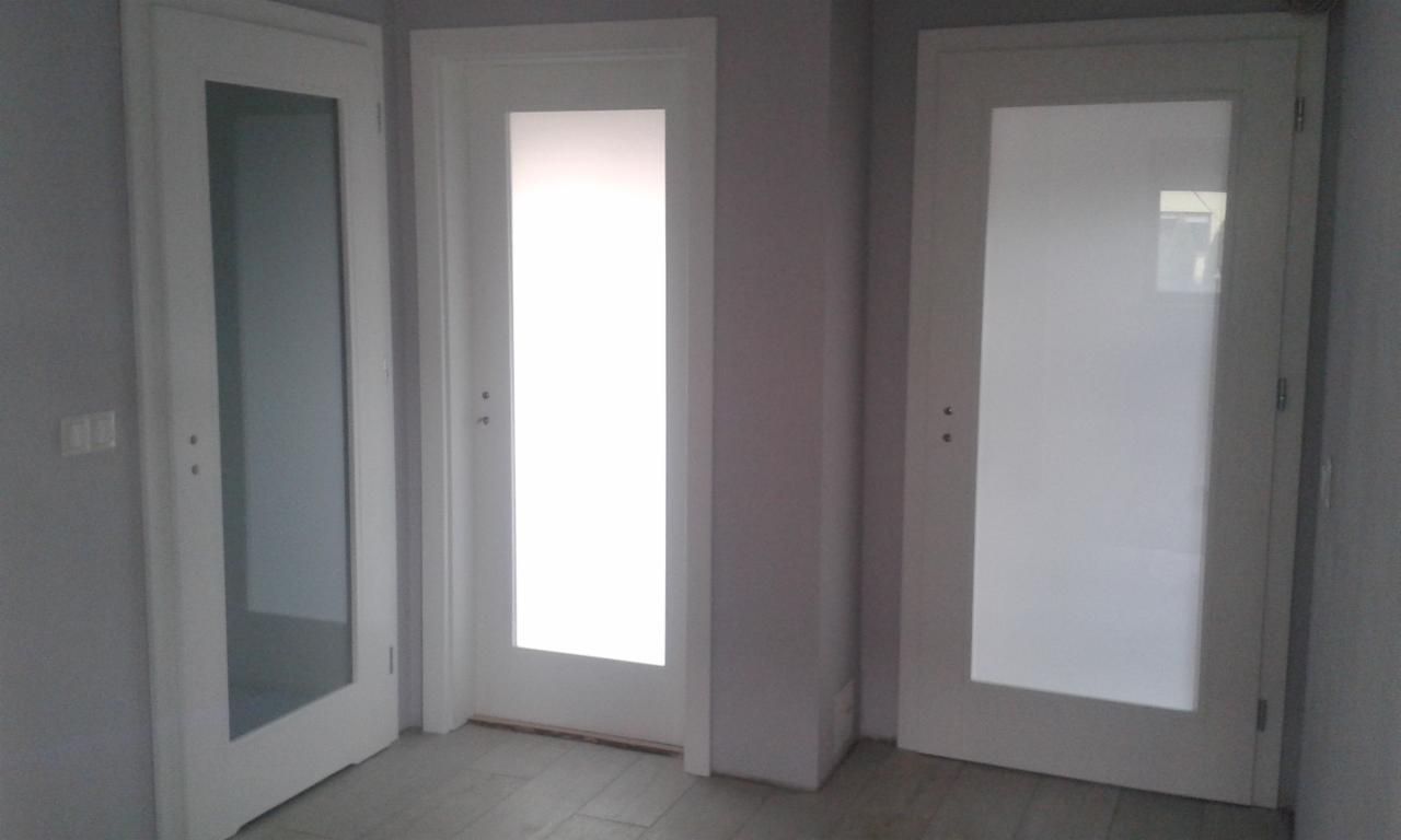 Trzy białe drzwi z przeszkleniami w różnych stylach, zamontowane w szarym pomieszczeniu z widoczną podłogą z płytek imitujących drewno.