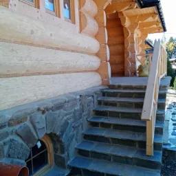 Zewnętrzne schody z ciemnego kamienia prowadzące do drewnianego domu z bali, widoczne kamienne obłożenie fundamentów i małe okienko piwniczne