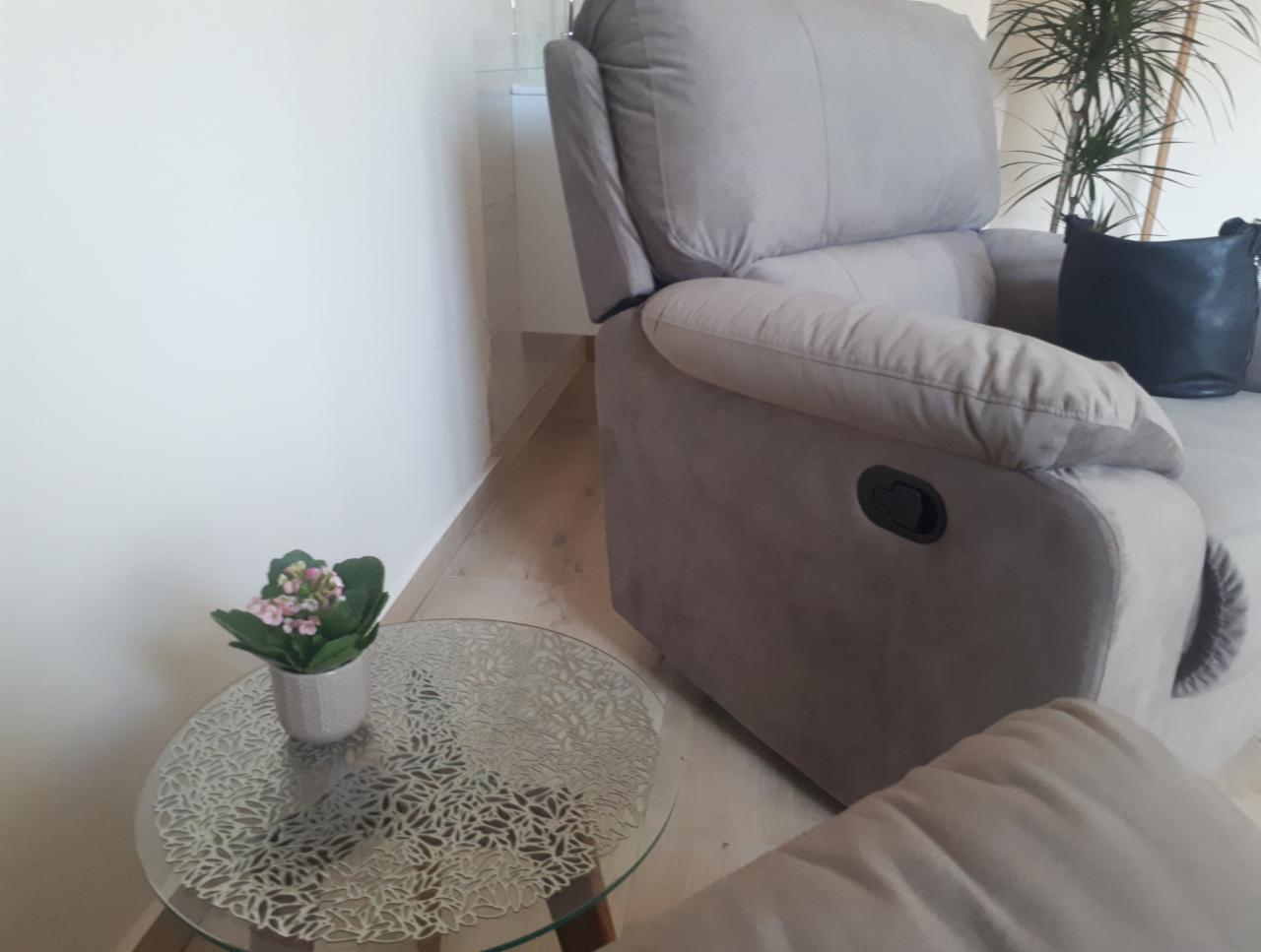 Jasnoszary fotel relaksacyjny z poduszką, obok okrągły stolik z ażurowym blatem i doniczką z drobnymi różowymi kwiatami, w tle roślina doniczkowa.