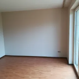 Puste pomieszczenie po remoncie z panelami podłogowymi w odcieniach brązu i beżu, świeżo pomalowanymi ścianami na jasny beż i białymi ramami okiennymi. Widoczne gniazdka elektryczne na ścianie.