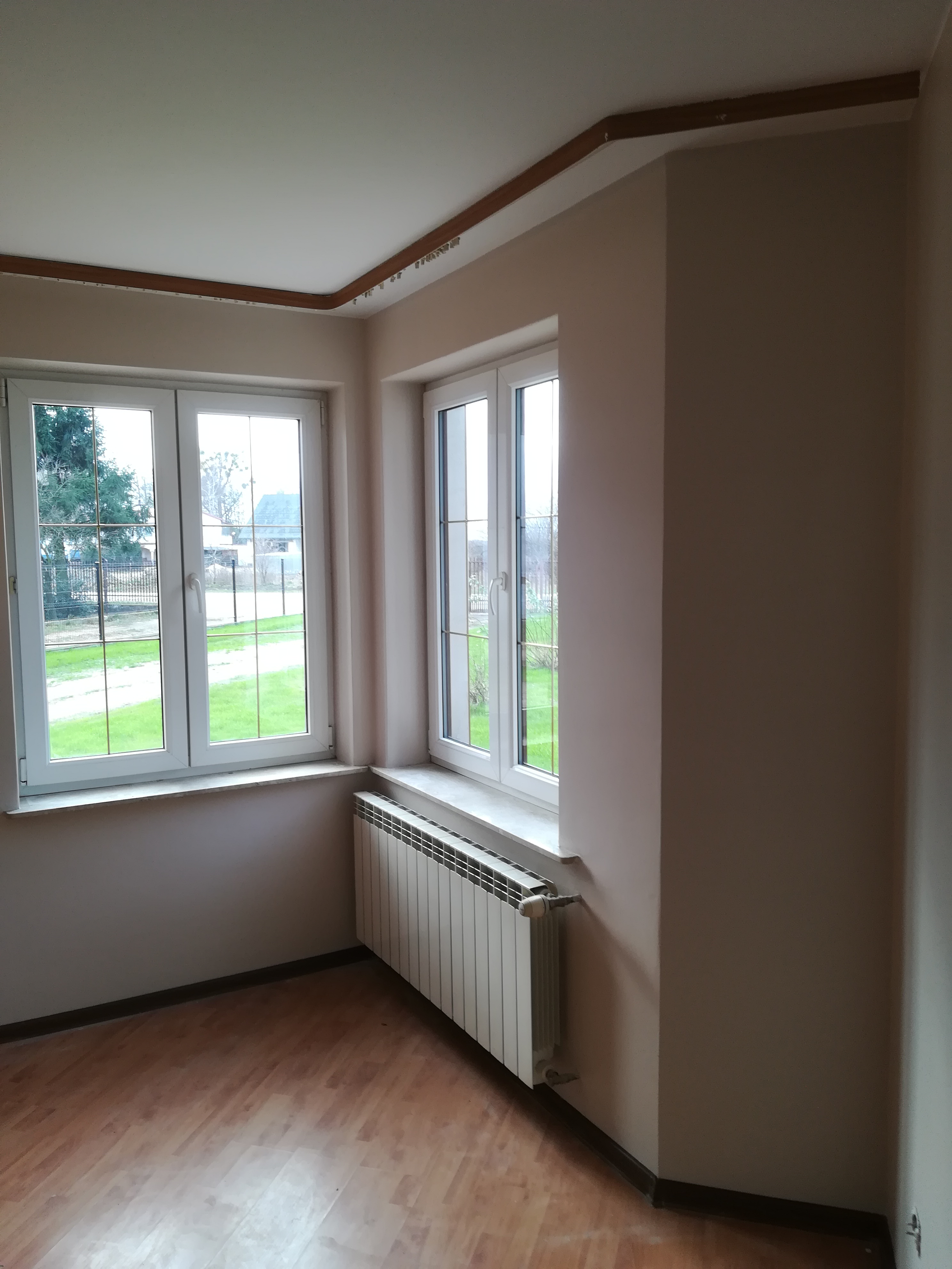 Jasny pokój po odświeżeniu, z dwoma oknami, białym grzejnikiem i drewnopodobną podłogą, ściany w odcieniach beżu, widoczne listwy sufitowe.