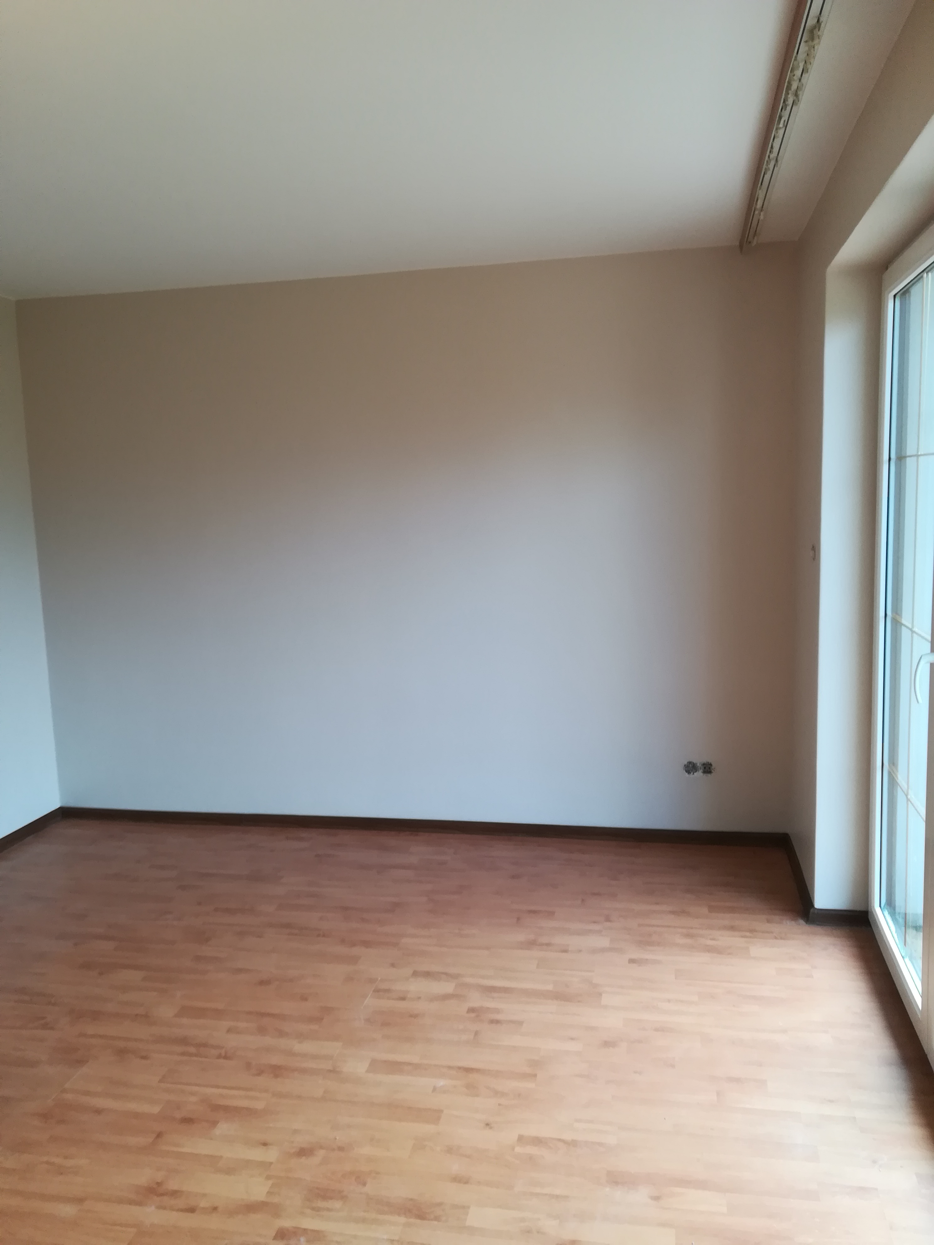 Puste pomieszczenie po remoncie z panelami podłogowymi w odcieniach brązu i beżu, świeżo pomalowanymi ścianami na jasny beż i białymi ramami okiennymi. Widoczne gniazdka elektryczne na ścianie.