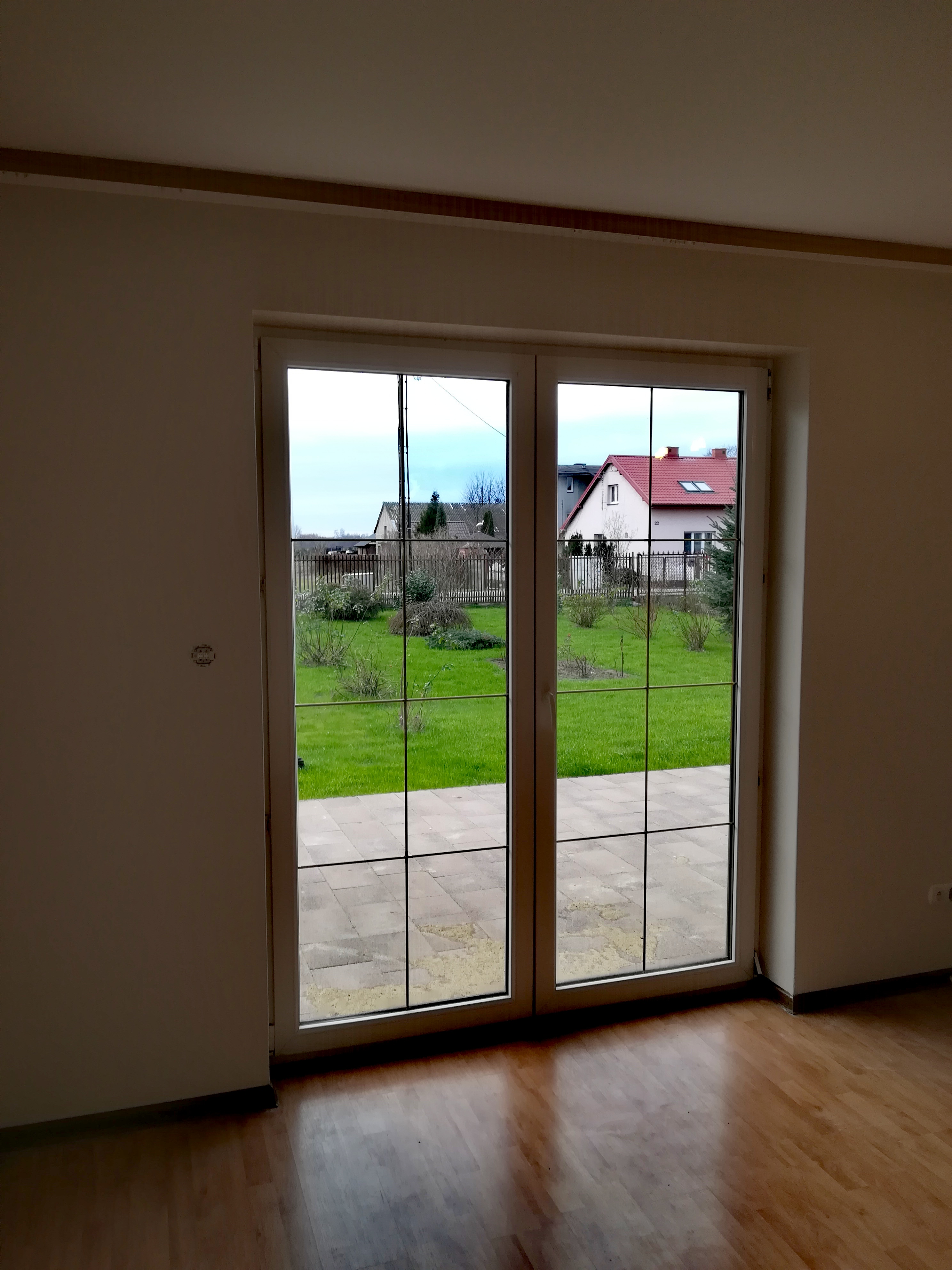 Puste, jasne pomieszczenie z parkietem, widok na ogród przez dwuskrzydłowe okno z szprosami. Ściany w odcieniach beżu, widoczne ślady po gniazdku elektrycznym. Ogród z trawnikiem i niskimi krzewami...