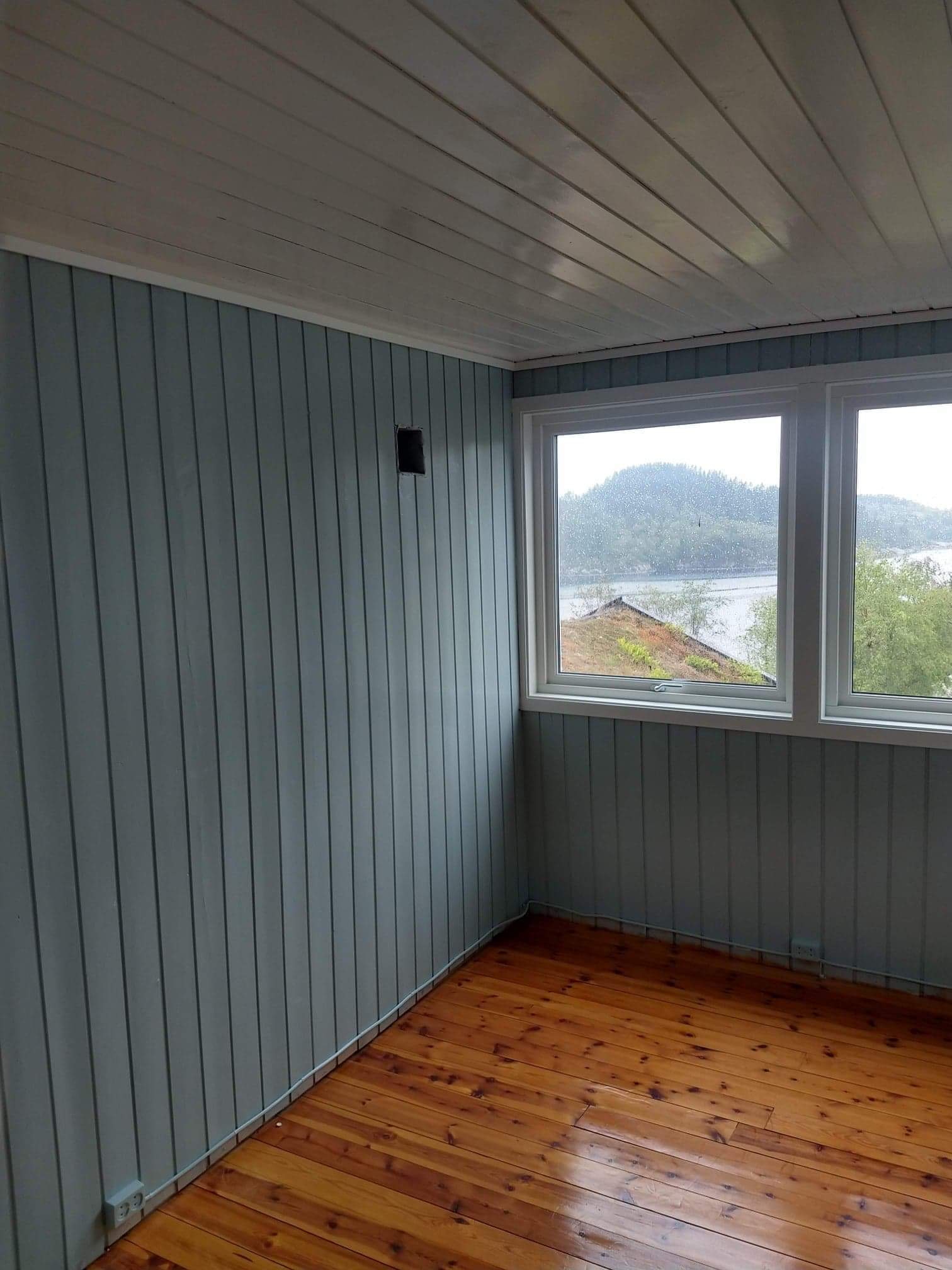 Wnętrze pokoju z drewnianą podłogą i pomalowanymi na szaro-niebiesko panelami ściennymi, widok na jezioro przez okno.