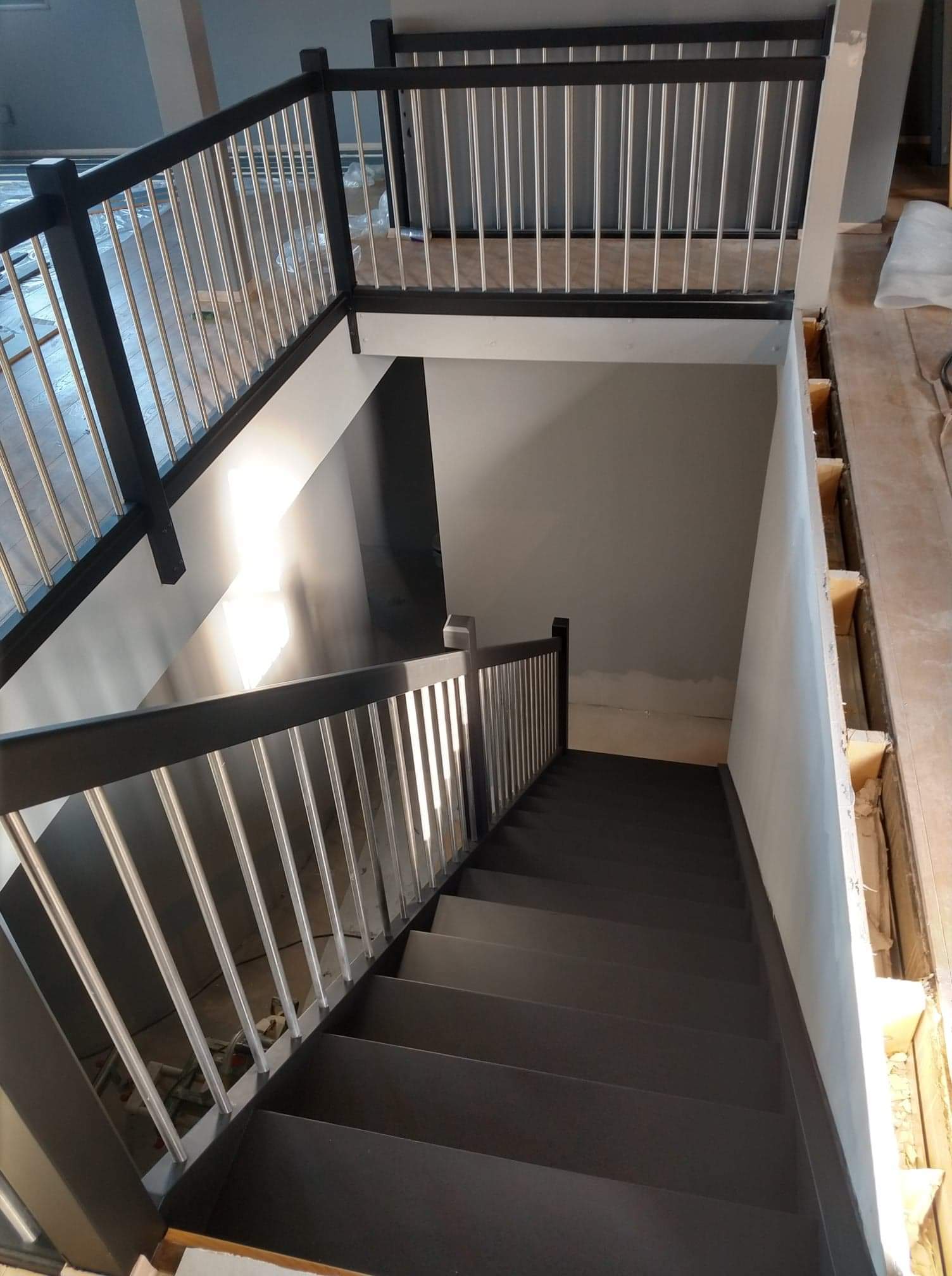 Nowoczesne schody z czarnymi stopniami i balustradą z metalowymi prętami, widoczne niedokończone wykończenie podłogi i ściany, jasne światło wpadające z boku.