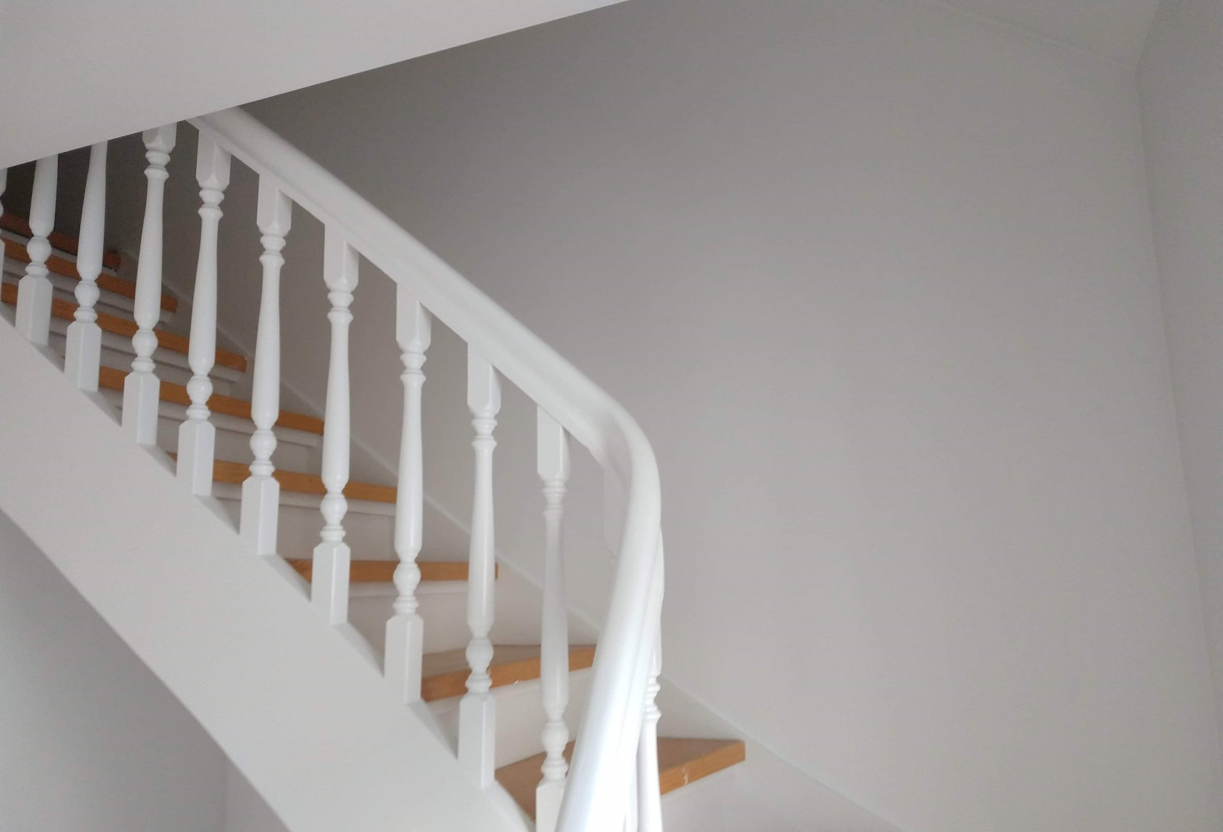 Świeżo odnowiona klatka schodowa z drewnianymi schodami i białą balustradą, ściany pomalowane na jasny odcień bieli.