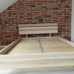 Rama łóżka z jasnego drewna z listwowym stelażem na tle ściany z cegły.