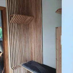 Drewniana zabudowa ściany z pionowych listew tworzących półkę i siedzisko z ciemną poduszką, obok szafka z drewna w jasnym odcieniu.