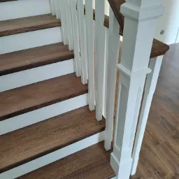 Widok z góry na fragment drewnianych schodów z białymi balustradami, gdzie widoczne są detale wykończenia i usłojenie drewna.