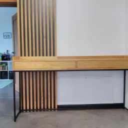 Nowoczesne biurko z blatem z jasnego drewna i czarną metalową ramą, z szufladami i dekoracyjną ścianką z pionowych listew drewnianych, widok na pokój dziecięcy w tle.