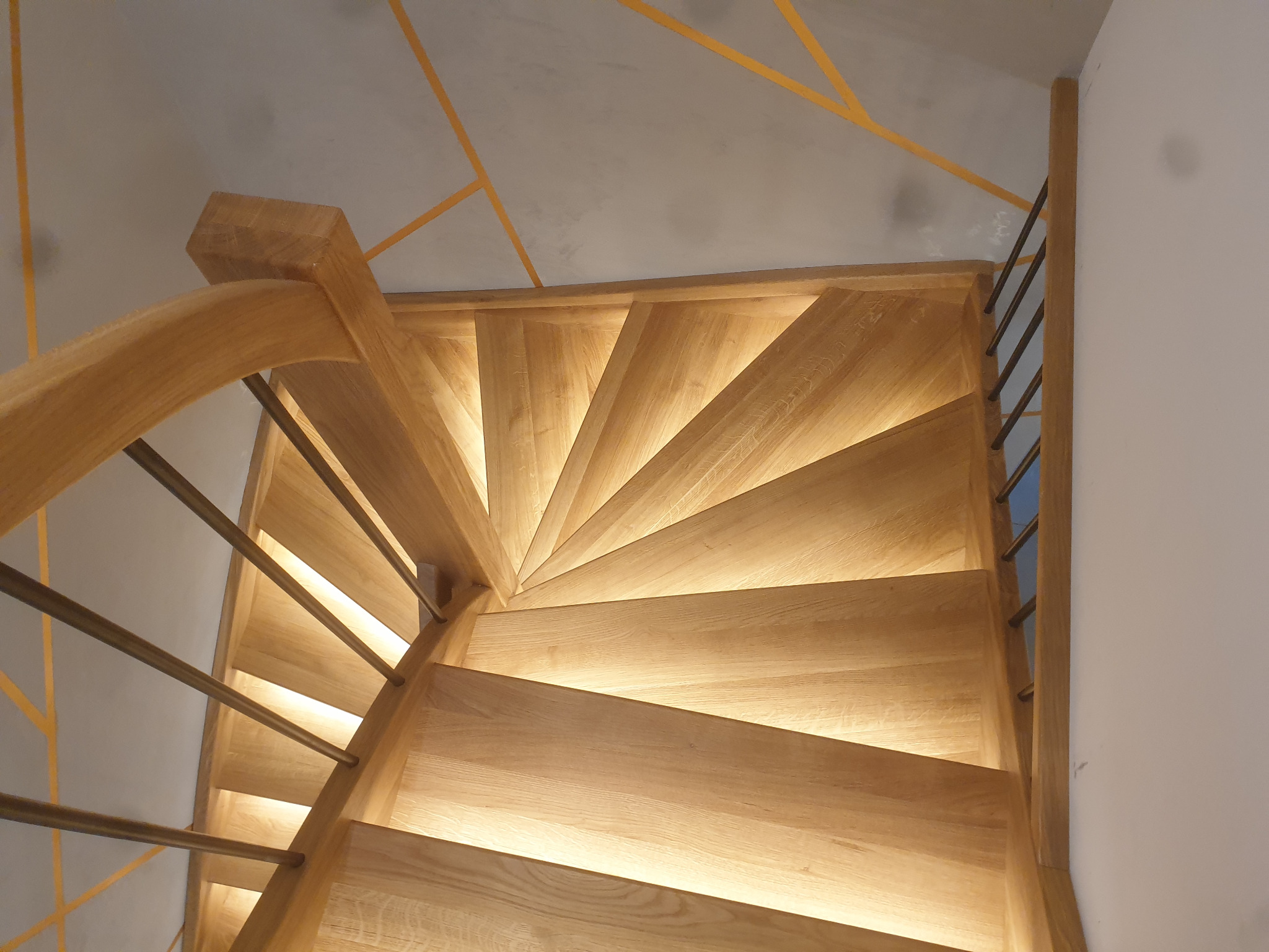 Spiralne stopnie z jasnego drewna z podświetleniem LED, widok z góry, metalowe barierki, sufit z geometrycznym wzorem w kolorze musztardowym.