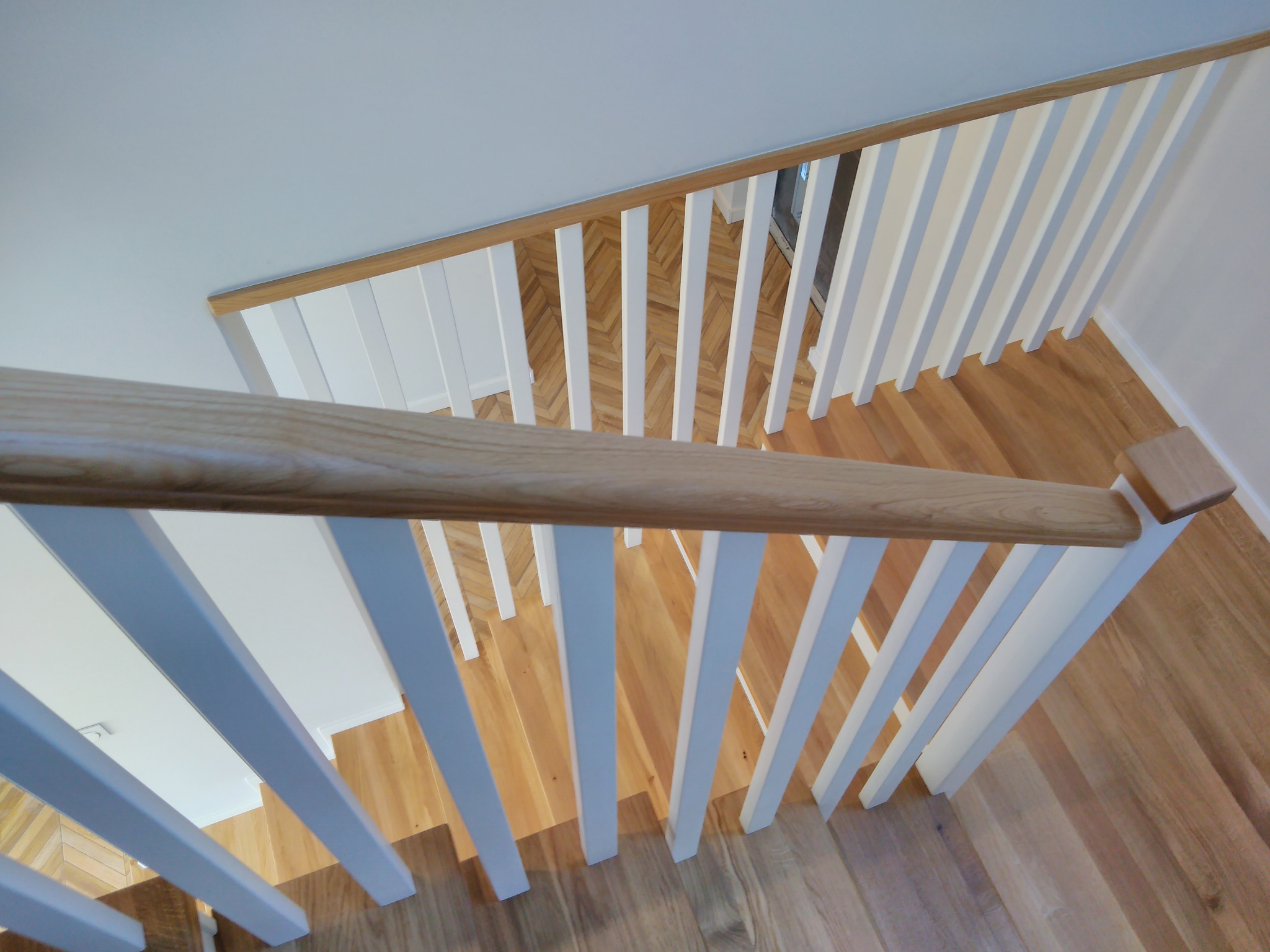 Widok z góry na jasne, drewniane schody z białymi balustradami, prowadzące na parkiet ułożony w jodełkę.