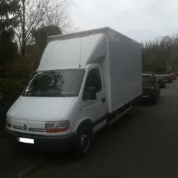 Biały samochód dostawczy Renault z wysoką zabudową skrzyniową, zaparkowany na chodniku z kostki, widok z przodu i boku, w tle drzewa i inne zaparkowane samochody.