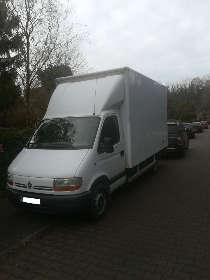 Biały samochód dostawczy Renault z wysoką zabudową skrzyniową, zaparkowany na chodniku z kostki, widok z przodu i boku, w tle drzewa i inne zaparkowane samochody.