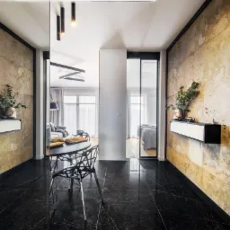 Nowoczesny korytarz z czarną marmurową podłogą, ścianami wyłożonymi kamieniem w odcieniach beżu i brązu, czarnym stolikiem z designerskim krzesłem oraz wiszącymi szafkami z dekoracjami roślinnymi.