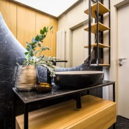 Nowoczesna łazienka z czarnym marmurem, okrągłym lustrem z podświetleniem, czarną umywalką nablatową i drewnianą szafką z otwartym regałem.