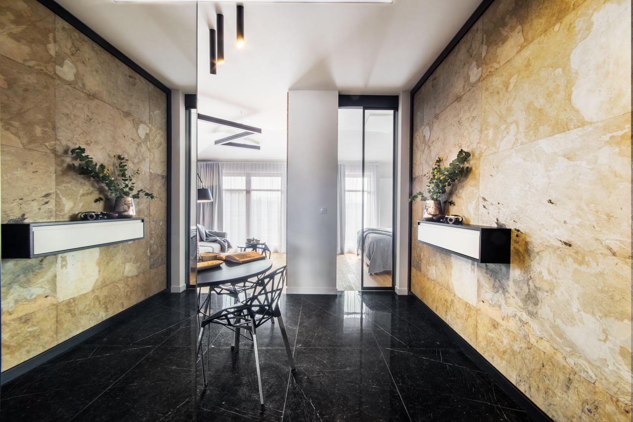 Nowoczesny korytarz z czarną marmurową podłogą, ścianami wyłożonymi kamieniem w odcieniach beżu i brązu, czarnym stolikiem z designerskim krzesłem oraz wiszącymi szafkami z dekoracjami roślinnymi.