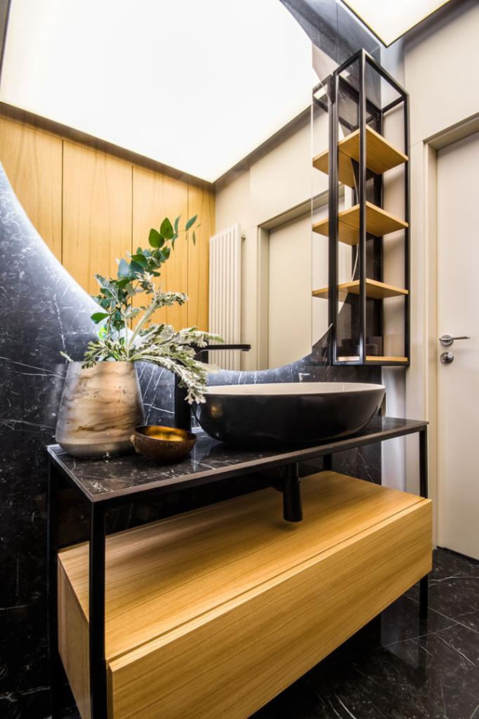 Nowoczesna łazienka z czarnym marmurem, okrągłym lustrem z podświetleniem, czarną umywalką nablatową i drewnianą szafką z otwartym regałem.