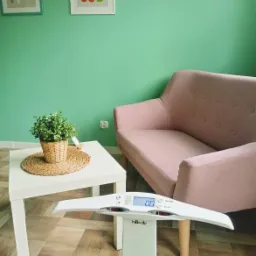 Wnętrze gabinetu z wagą InBody, stolikiem z rośliną w doniczce i pastelową kanapą na tle miętowej ściany z obrazkami owoc&oacute;w i zi&oacute;ł.