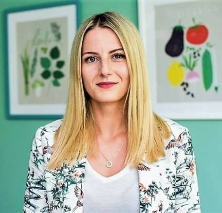 Portret kobiety z blond włosami w kwiecistej marynarce, na tle jasnozielonej ściany z dwoma oprawionymi grafikami z motywami roślinnymi i warzywnymi.