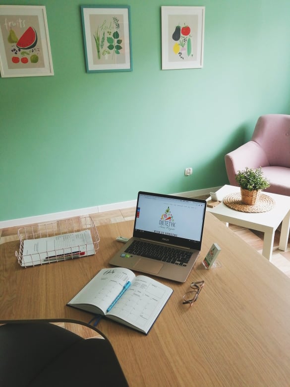 Biurko z otwartym kalendarzem, laptopem wyświetlającym stronę z logo, okularami i metalowym koszykiem na dokumenty; w tle zielona ściana z trzema grafikami owoców i warzyw oraz różowy fotel...