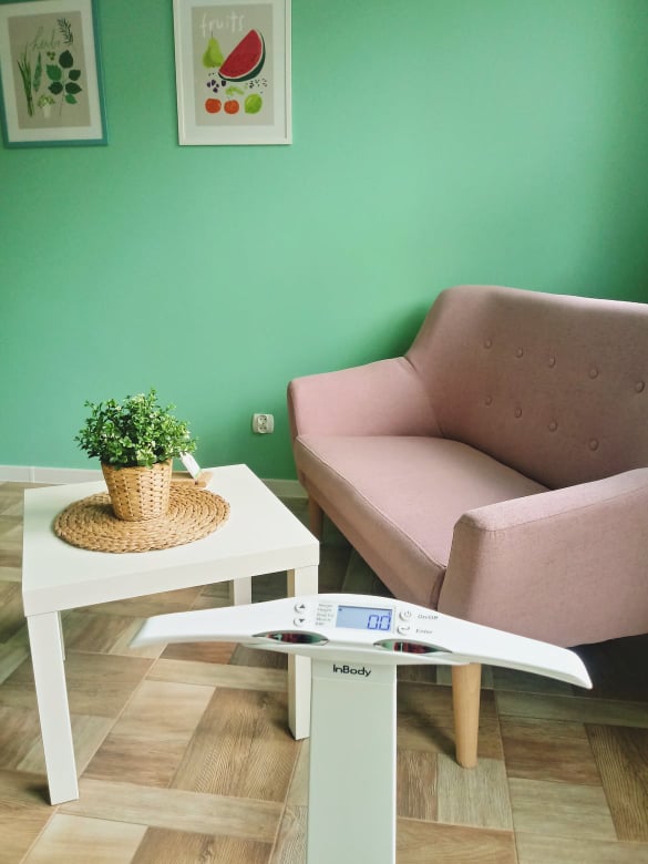 Wnętrze gabinetu z wagą InBody, stolikiem z rośliną w doniczce i pastelową kanapą na tle miętowej ściany z obrazkami owoców i ziół.