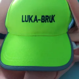 Jasnozielona czapka z daszkiem z haftowanym napisem 'LUKA-BRUK' firmy z Brzezin, leżąca na różowej odzieży.