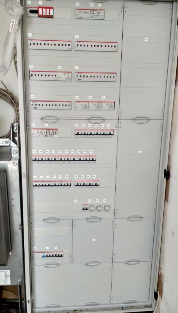 Rozdzielnica elektryczna z wieloma zabezpieczeniami i wyłącznikami, zamontowana w szarej obudowie, widok z bliska.