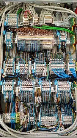 Rozdzielnica elektryczna z licznymi przewodami i złączkami, widoczne kable w kolorach niebieskim, brązowym i zielonym, ułożone w uporządkowany sposób.