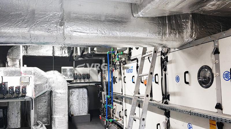 Instalacja centrali wentylacyjnej w pomieszczeniu technicznym szpitala, widoczne izolowane przewody wentylacyjne pod sufitem, drabina oparta o centralę oraz rury z zaworami.