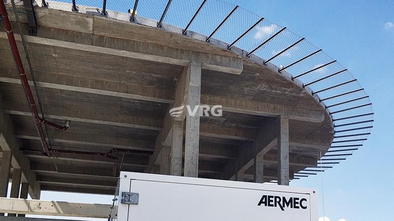 Szpital Miejski w Zabrzu: Widok z dołu na surową betonową konstrukcję stropu z widoczną instalacją rur i siatką zabezpieczającą na obrzeżach, z białym agregatem marki Aermec w dolnej części kadru.