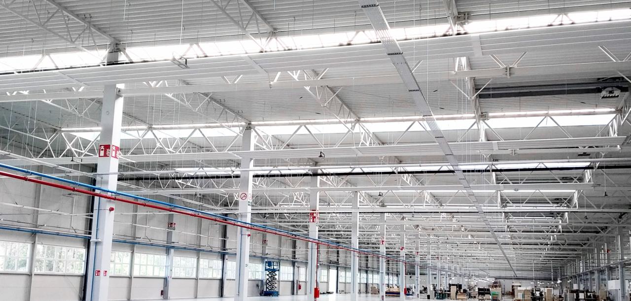 Widok wnętrza hali przemysłowej z widocznymi elementami konstrukcji stalowej, instalacjami wentylacyjnymi z rurami w kolorach czerwonym i niebieskim, oraz oknami w dolnej części ściany.