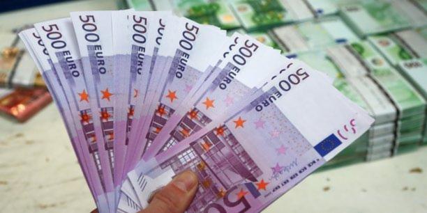 Dłoń trzymająca wachlarz banknotów 500 euro, w tle stosy banknotów 100 euro.
