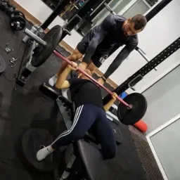 Kobieta wyciska sztangę na ławce pod okiem mężczyzny w siłowni, lustrzane odbicie w tle