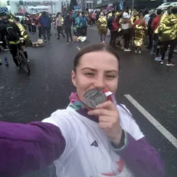 Uśmiechnięta kobieta robi selfie z medalem na szyi po biegu ulicznym, w tle widoczni inni uczestnicy w złotych płaszczach termicznych i brama startowa.