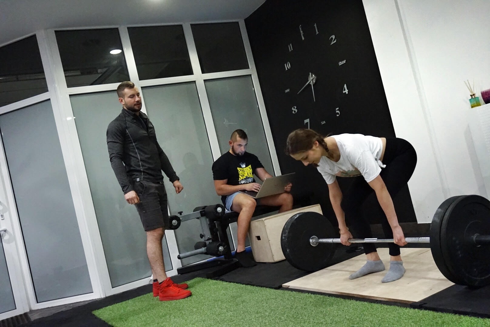 Kobieta w Rzeszowie przygotowuje się do podnoszenia sztangi pod okiem trenera, podczas gdy drugi mężczyzna siedzi z laptopem, analizując dane treningowe.