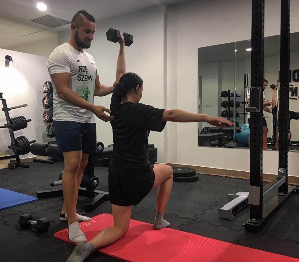 Trener personalny asystuje kobiecie w ćwiczeniu z hantlą w pozycji klęczącej na macie; w tle lustro i sprzęt do ćwiczeń.