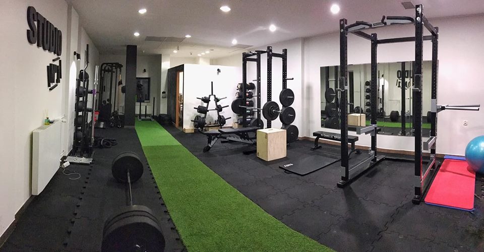 Wnętrze przestronnej siłowni z czarną podłogą z puzzli, sztuczną trawą, sprzętem do ćwiczeń siłowych, lustrem i napisem 'Studio' na ścianie.