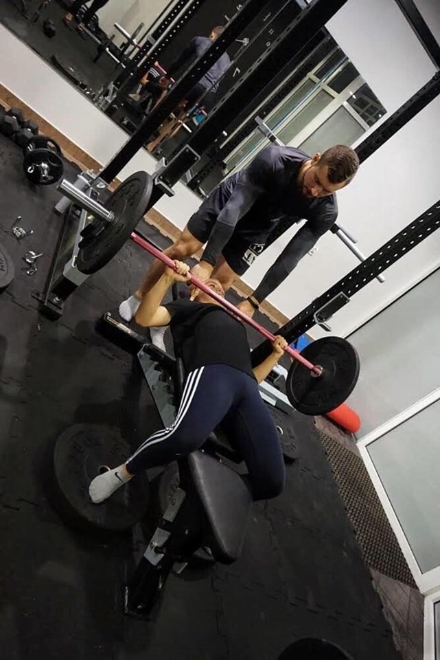 Kobieta wyciska sztangę na ławce pod okiem mężczyzny w siłowni, lustrzane odbicie w tle