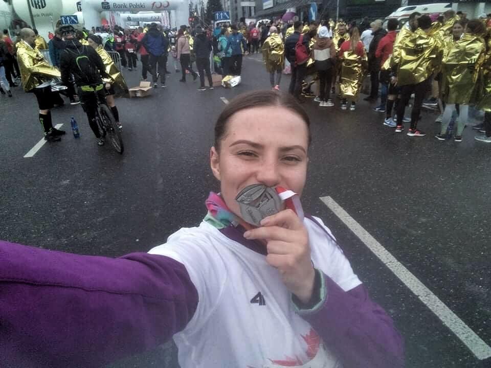 Uśmiechnięta kobieta robi selfie z medalem na szyi po biegu ulicznym, w tle widoczni inni uczestnicy w złotych płaszczach termicznych i brama startowa.