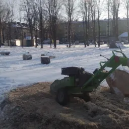 Pracownik w odblaskowej kurtce obsługuje frezarkę do pni drzew na ośnieżonym placu miejskim, w tle ścięte pnie i budynki.