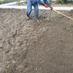 Przygotowywanie ziemi pod trawnik: ogrodnik w niebieskich jeansach i czerwonych rękawicach grabi ziemię, widoczne ślady stóp na ciemnej, wilgotnej glebie.