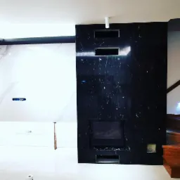 Częściowo wykończona ściana z czarnego kamienia z wbudowanym kominkiem i otworami wentylacyjnymi, sąsiadująca z białą ścianą z imitacją marmuru, widoczne fragmenty schodów.