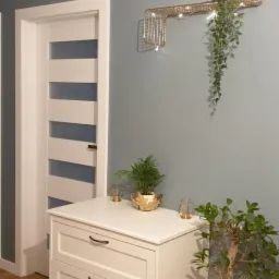 Korytarz z komodą w stylu klasycznym, białe drzwi z przeszkleniami, dekoracyjna metalowa półka ścienna z rośliną zwisającą, ściana w kolorze gołębim, podłoga drewniana.