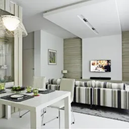 Jasne, nowocześnie urządzone wnętrze salonu z jadalnią, z białym stołem i krzesłami oraz sofą w paski, oświetlone lampą z ażurowym abażurem. Na suficie widoczne podłużne, wpuszczane oprawy...