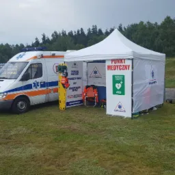Ambulans i namiot punktu medycznego AlfaMedyk na trawiastym terenie, widoczna deska ortopedyczna i krzesełko ratownicze, w tle trybuny i las.